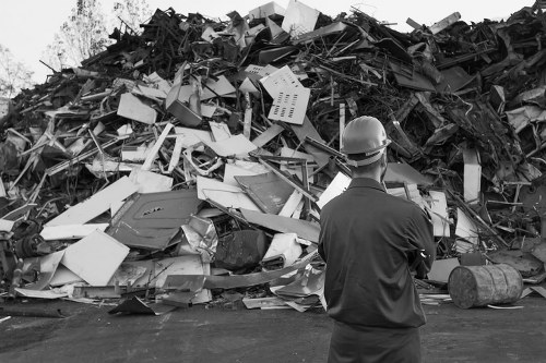 A man wearing a safety helmet and dark jacket stands with his back to the camera in front of a large pile of mixed construction and industrial waste on a paved surface, likely outside a waste disposal or recycling facility. The waste heap contains various materials such as broken plastic items, foam, metal fragments, wooden planks, and discarded appliances, all in a disordered arrangement. Some objects are stacked haphazardly, with sharp edges and rough textures visible. In the background, the environment appears industrial, with no specific building features visible, indicating an outdoor waste storage area common in commercial rubbish removal services. The scene is depicted in black and white, emphasizing the volume and disorder of the waste, aligning with the context of professional rubbish clearance often undertaken by companies such as Business Waste Removal Barking, serving local areas including Barking and the surrounding postcode districts.