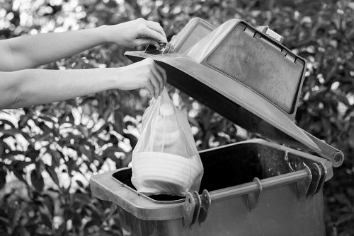 A person is lifting the lid of a grey waste bin outdoors, revealing a plastic bag filled with assorted rubbish including white bowls or containers. The waste bin is positioned on a paved surface, with the lid opened fully, exposing the black interior of the bin. The background features leafy vegetation, indicating a garden or courtyard setting. The scene is captured in monochrome, emphasizing the textures of the plastic bag, the bin's smooth surface, and the surrounding foliage. This image illustrates typical rubbish disposal activities, aligning with the waste removal services offered by Business Waste Removal Barking in the local area. The presence of the waste bin on the pavement suggests regular waste collection in the Barking district, part of the provider's professional rubbish management solutions.
