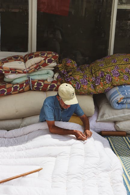 Inside a room with large glass windows, an elderly person wearing a beige cap, a blue shirt, and white sleeves is seated on the edge of a white mattress, folding or inspecting fabric. Behind them, the