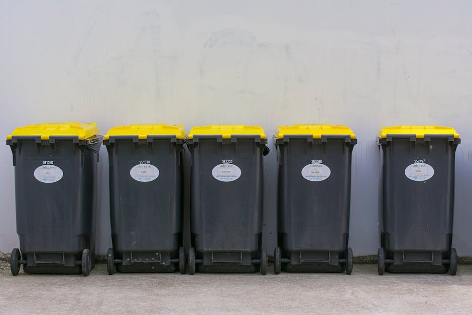 A row of large, rectangular wheelie bins lined up outdoors along a paved pathway next to a brick wall. The bins are made of hard plastic with textured surfaces on their lids, each fitted with handles 